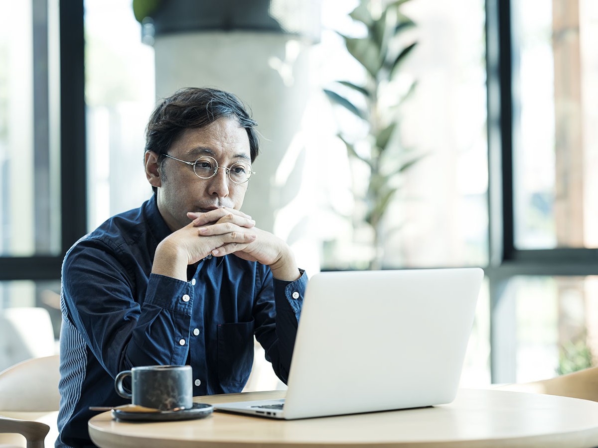 Man with glasses in a blue shirt sitting at a table with coffee looking at his laptop.