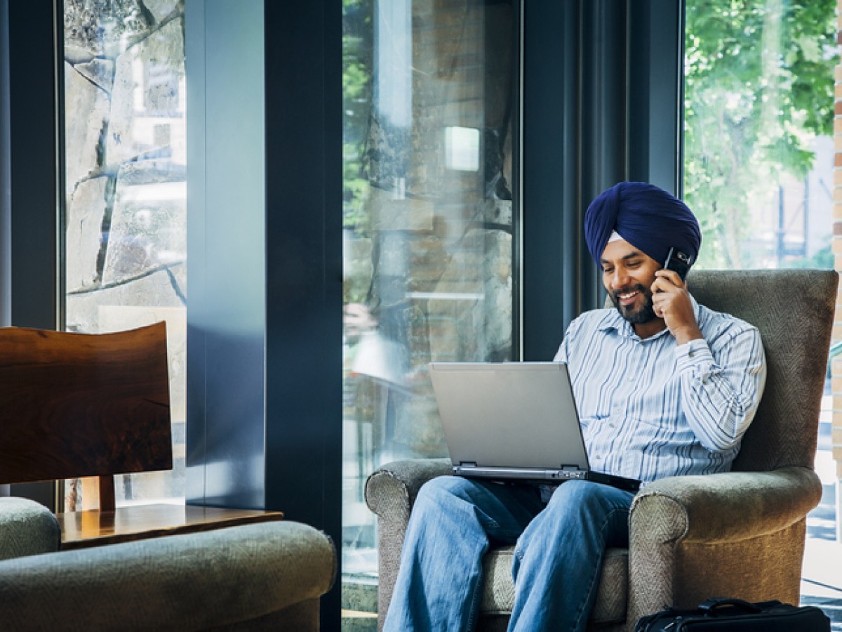 A smiling South Asian man using a laptop at home, while talking on a cell phone