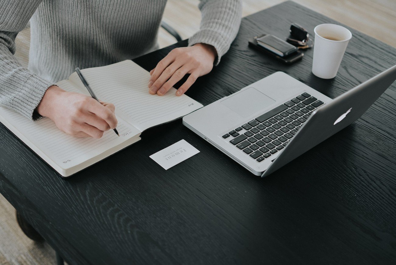 Desk with laptop and woman's hands writing in a notebook.