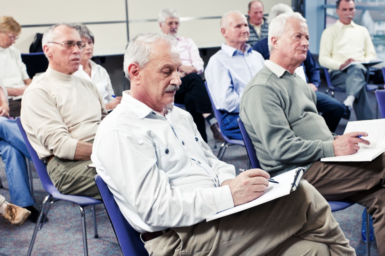 A room full of seniors, mostly men, seated in rows at a class or seminar