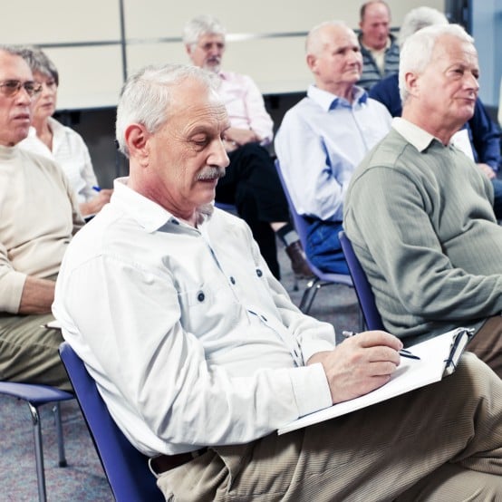 A room full of seniors, mostly men, seated in rows at a class or seminar